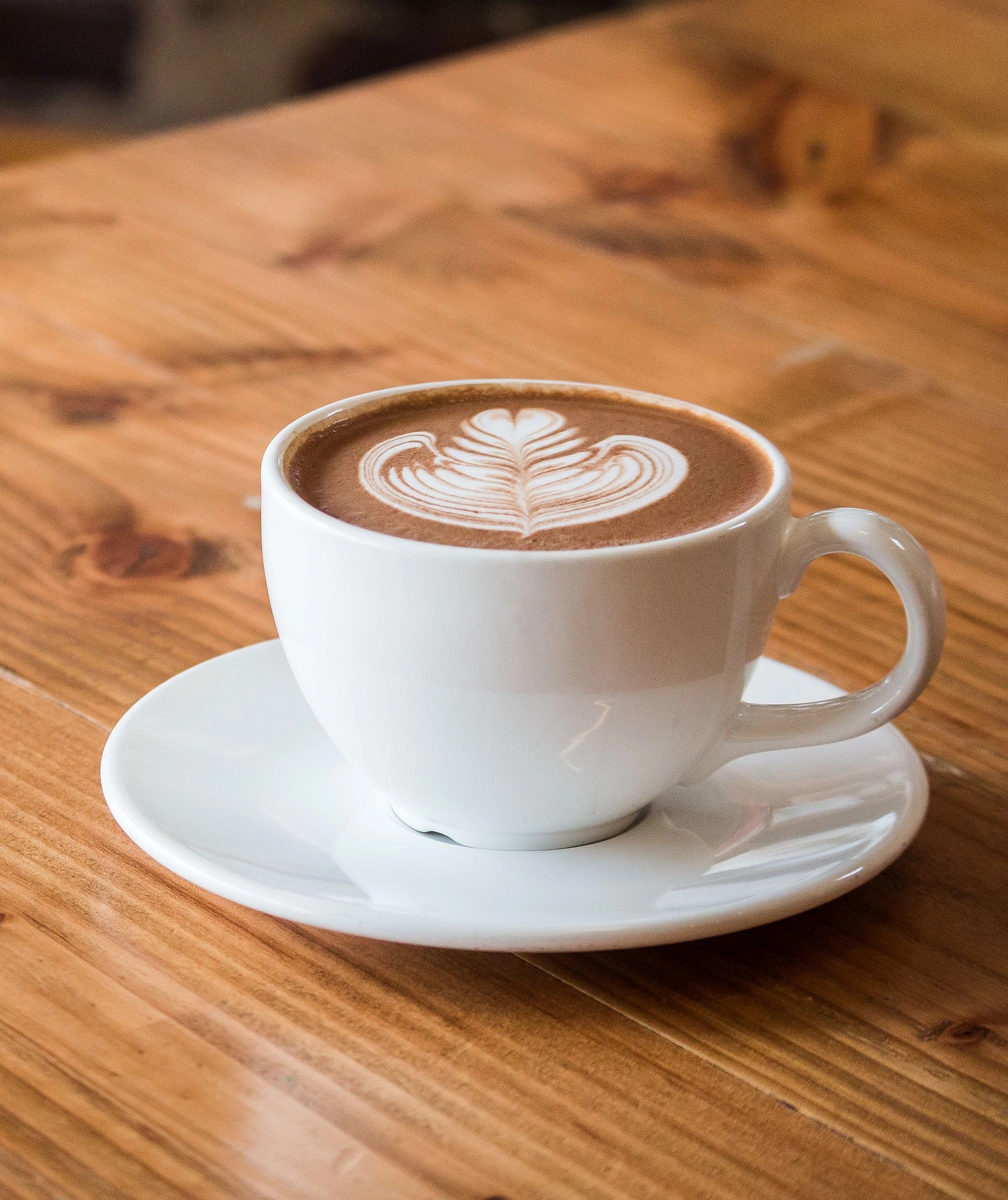 A complete white ceramic cup of coffee with leaf-shaped latte art, its handle and saucer visible on a warm wooden table